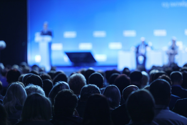 Publikum bei einer Konferenz mit verschwommenen Sprechern auf einer blau beleuchteten Bühne. - © Gennady Danilkin - stock.adobe.com