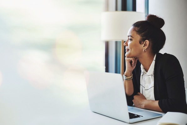 Eine Frau mit Dutt sitzt an einem Laptop und blickt nachdenklich aus dem Fenster eines hellen Raums. - © Valerie M/peopleimages.com - stock.adobe.com